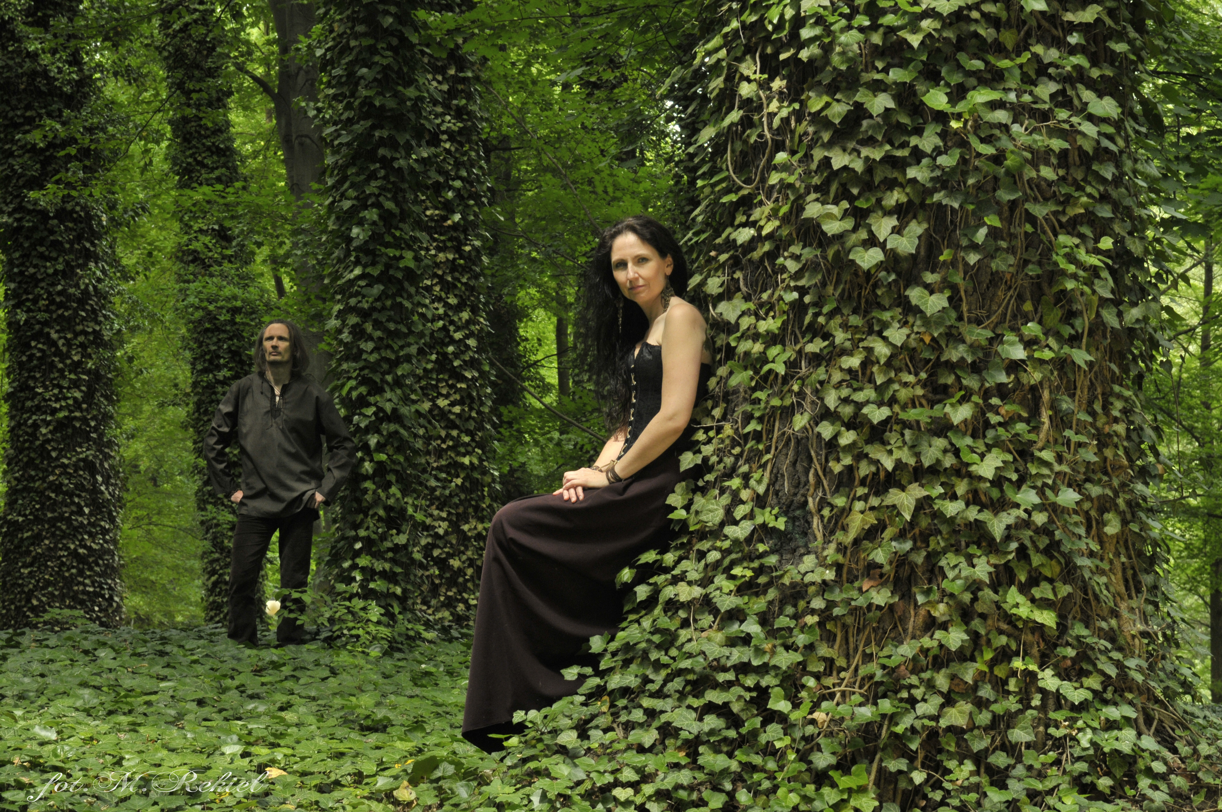 Kobieta (Anna Winiarska) ubrana na czarno opiera się  o drzewo. W oddali stoi mężczyzna ubrany na czarno. Drzewa porośnięte są bluszczem